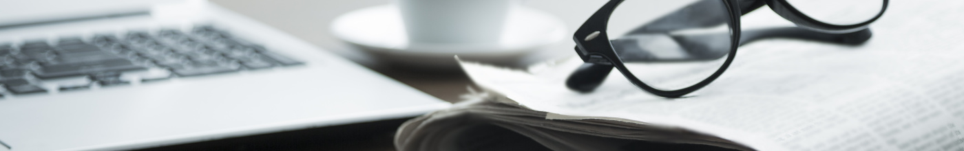 An image of a laptop, newspaper, reading glasses, coffee cup, and a pen on a desk.