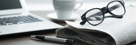 An image of a laptop, newspaper, reading glasses, coffee cup, and a pen on a desk.